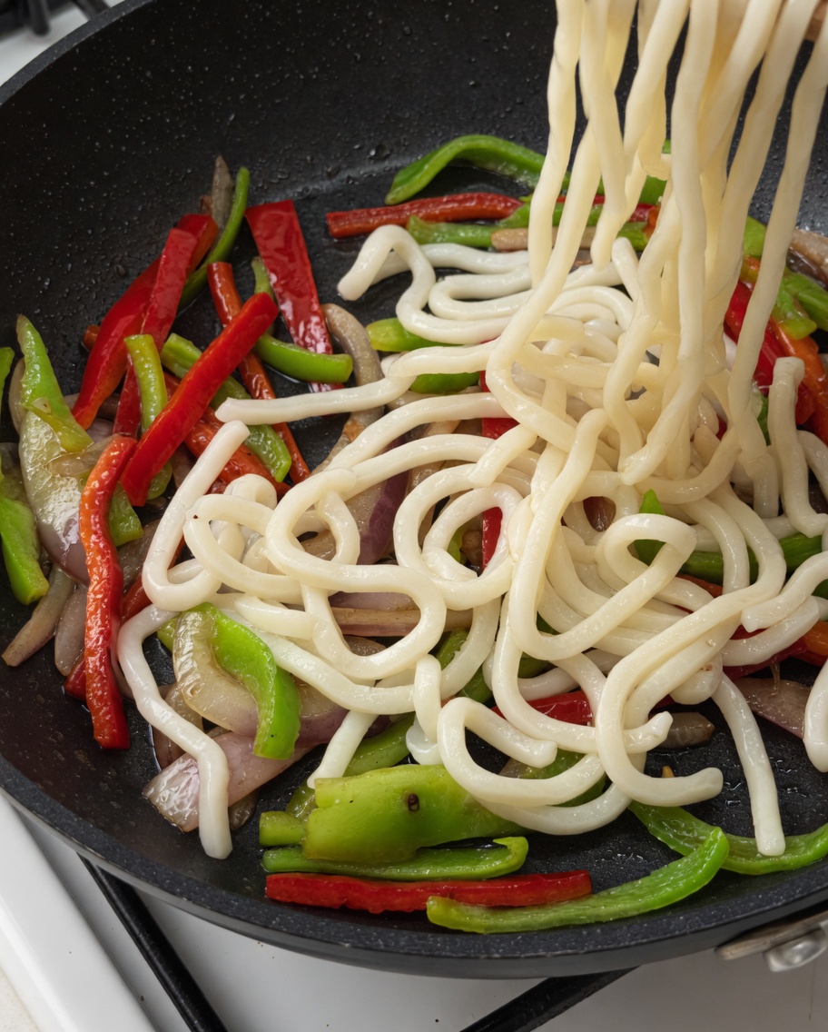 Nouilles udon et lanières de bœuf ajoutées dans la poêle contenant les légumes.