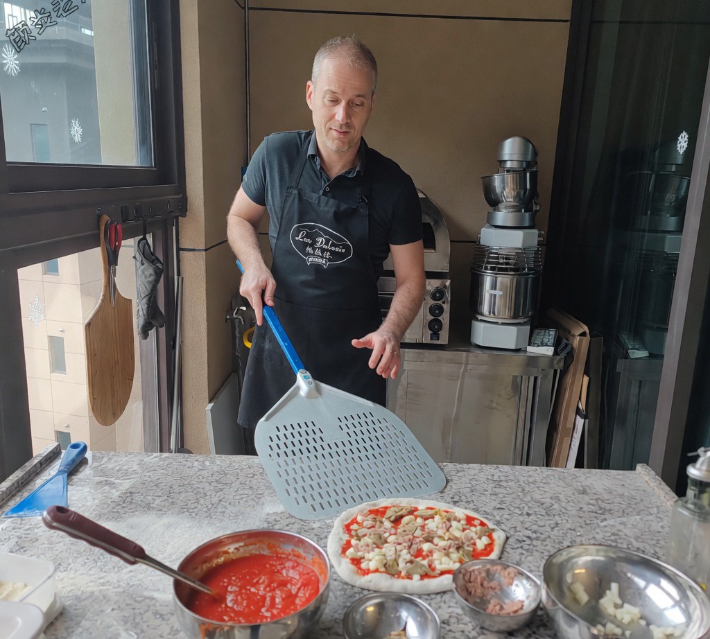 Une pelle à pizza en métal glissée sous la pizza crue assemblée pour la préparer à la cuisson.