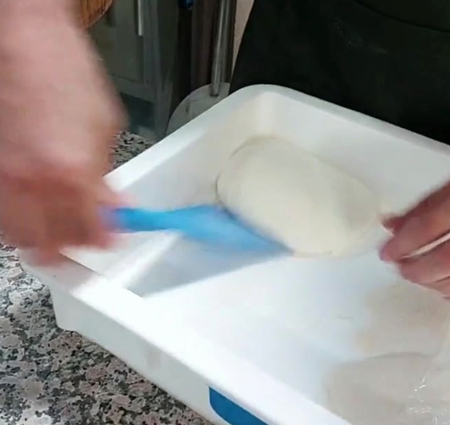 Une portion de pâte à pizza soulevée dun récipient avec un grattoir en plastique bleu.