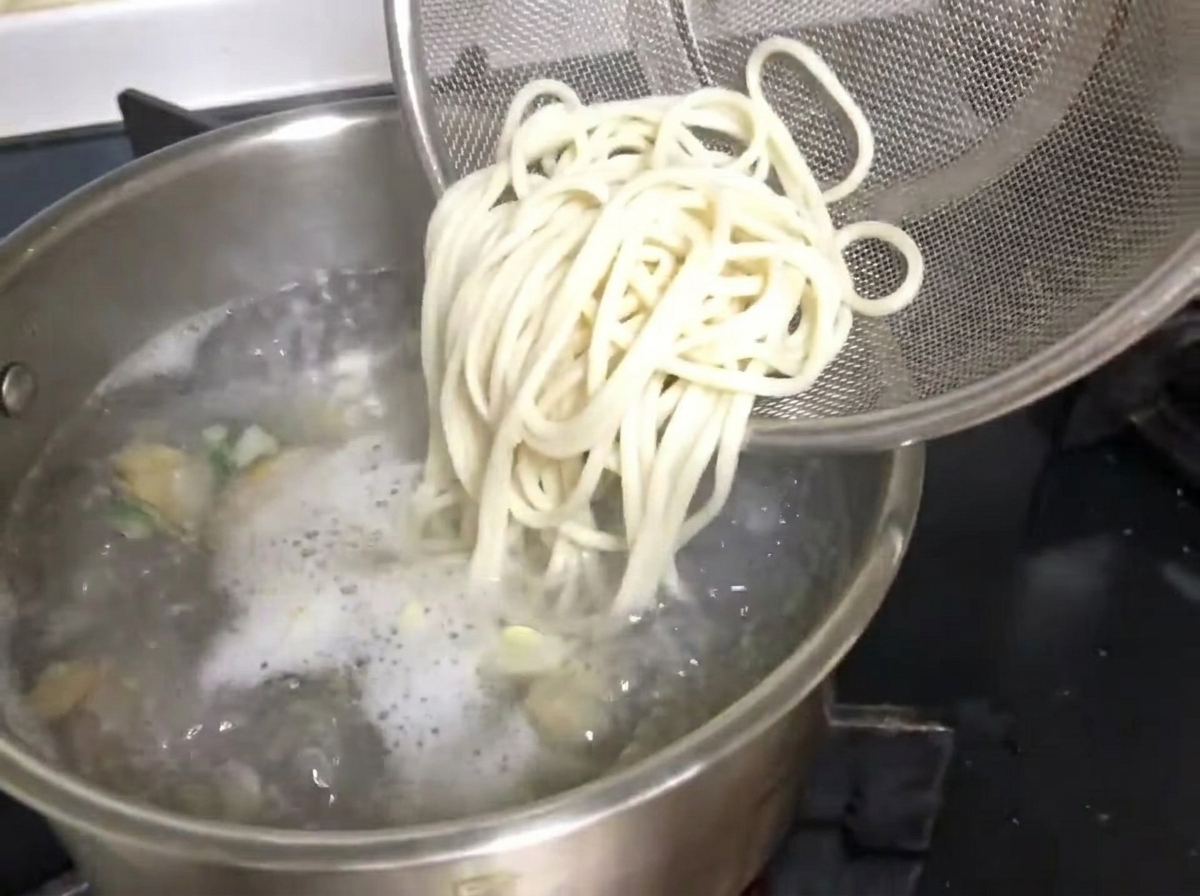 Des nouilles rincées glissant dune passoire dans une marmite de bouillon de palourdes en ébullition.