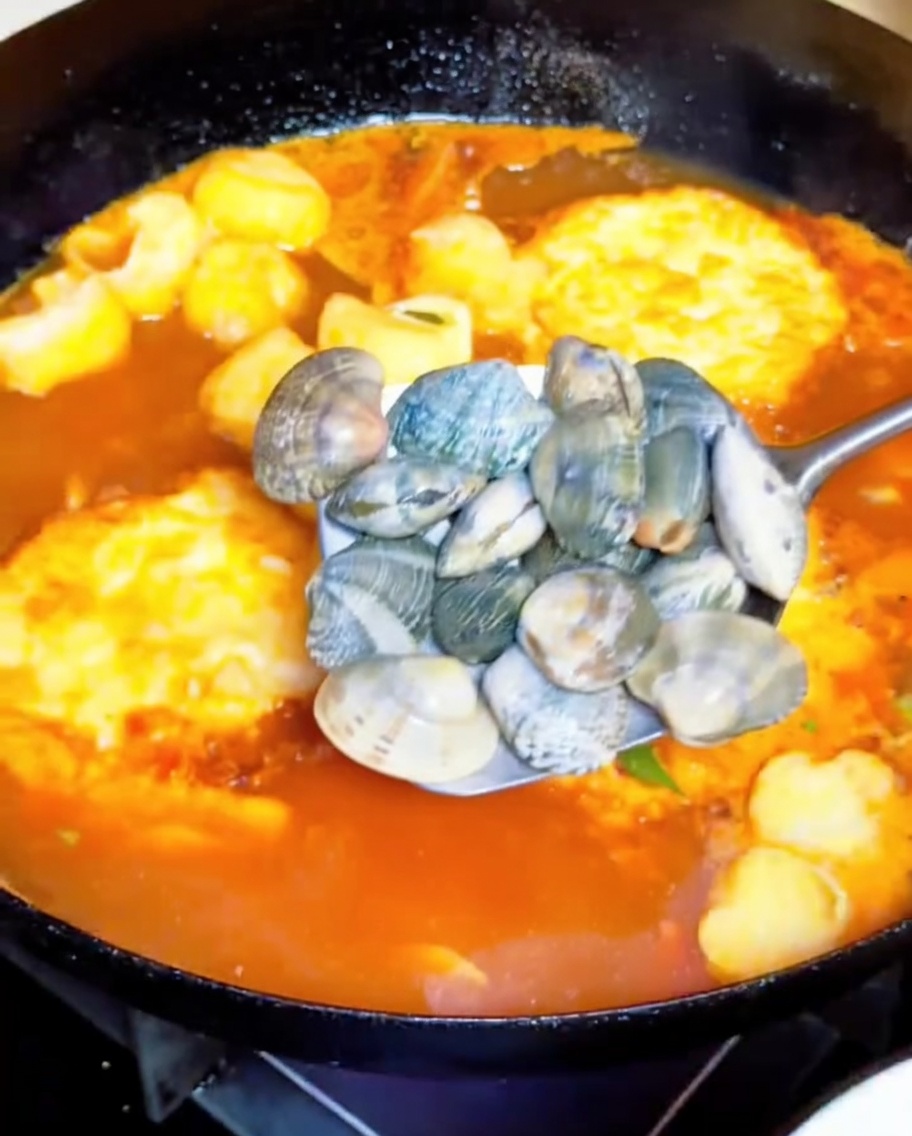 Une louche en métal ajoutant des palourdes entières dans un bouillon de tomates bouillonnant.