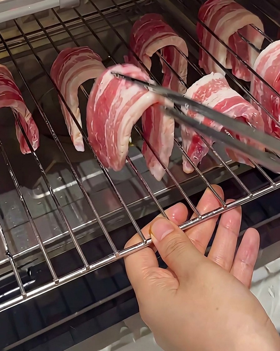 Tranches de poitrine de porc crue suspendues sur les fils dune grille de four, tenues par des pinces.
