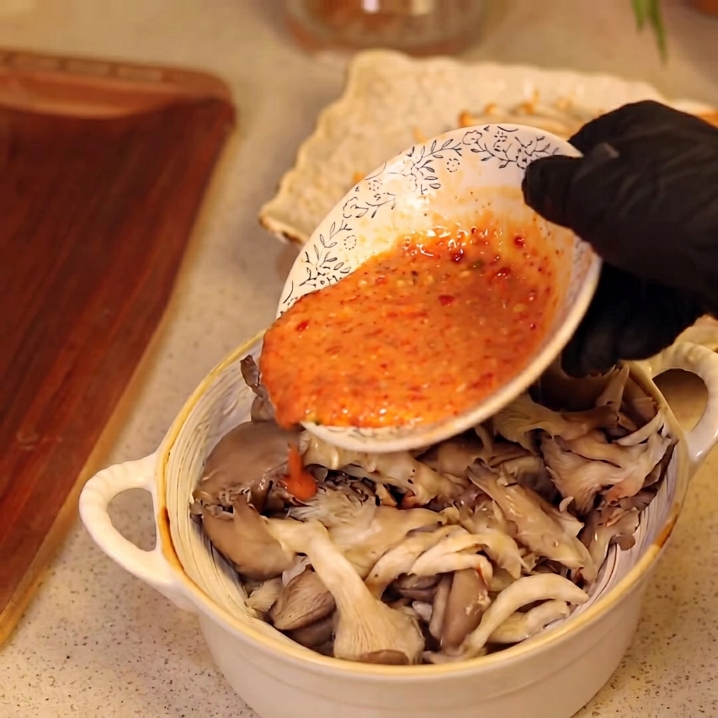 Une main gantée versant une sauce sur un bol de champignons.