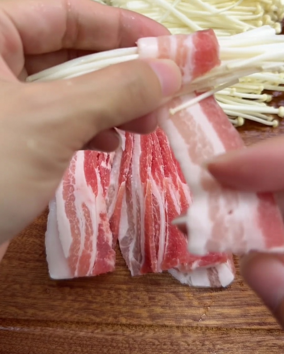 Mains enroulant une fine tranche de poitrine de porc autour dun petit bouquet de champignons enoki.
