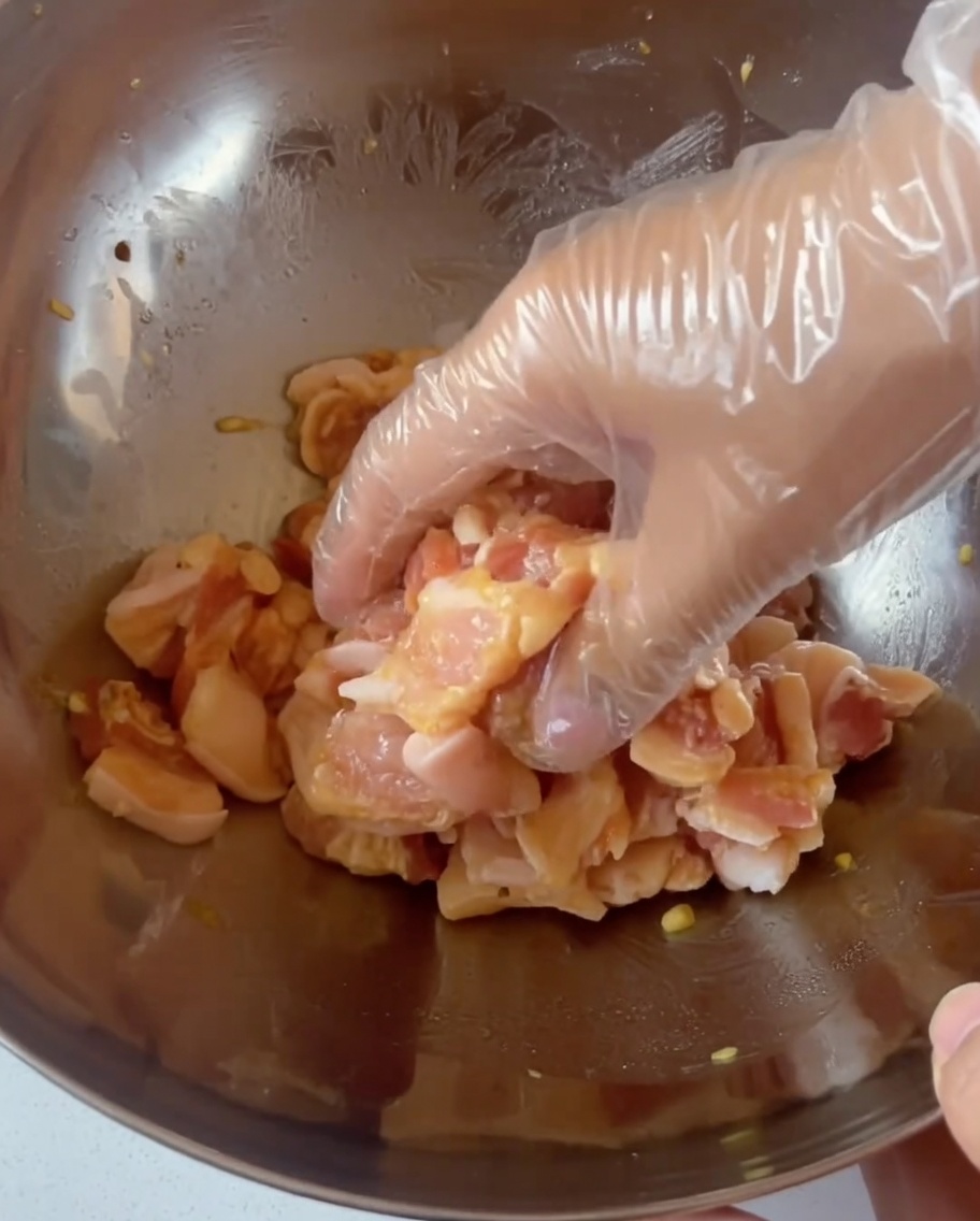 Une main gantée mélangeant des tranches de poitrine de porc assaisonnées dans un grand bol en acier inoxydable.