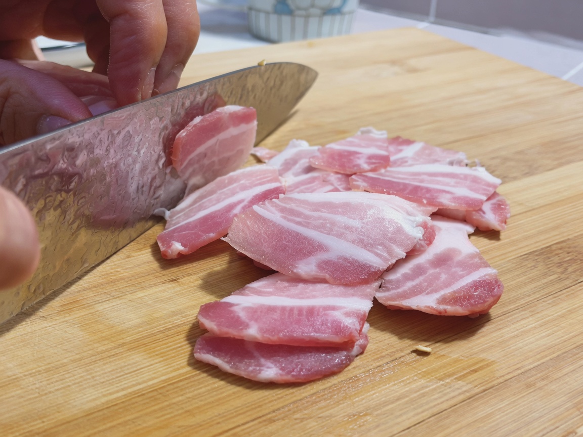 Fines tranches de poitrine de porc crue disposées sur une planche à découper en bois à côté dun grand couteau de chef.