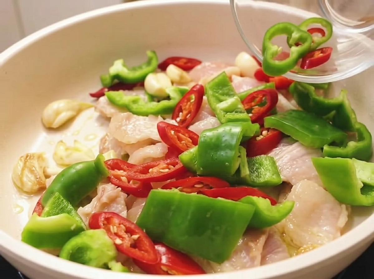 Poivrons rouges et verts fraîchement hachés versés depuis un petit bol en verre dans une poêle de porc tranché partiellement cuit.