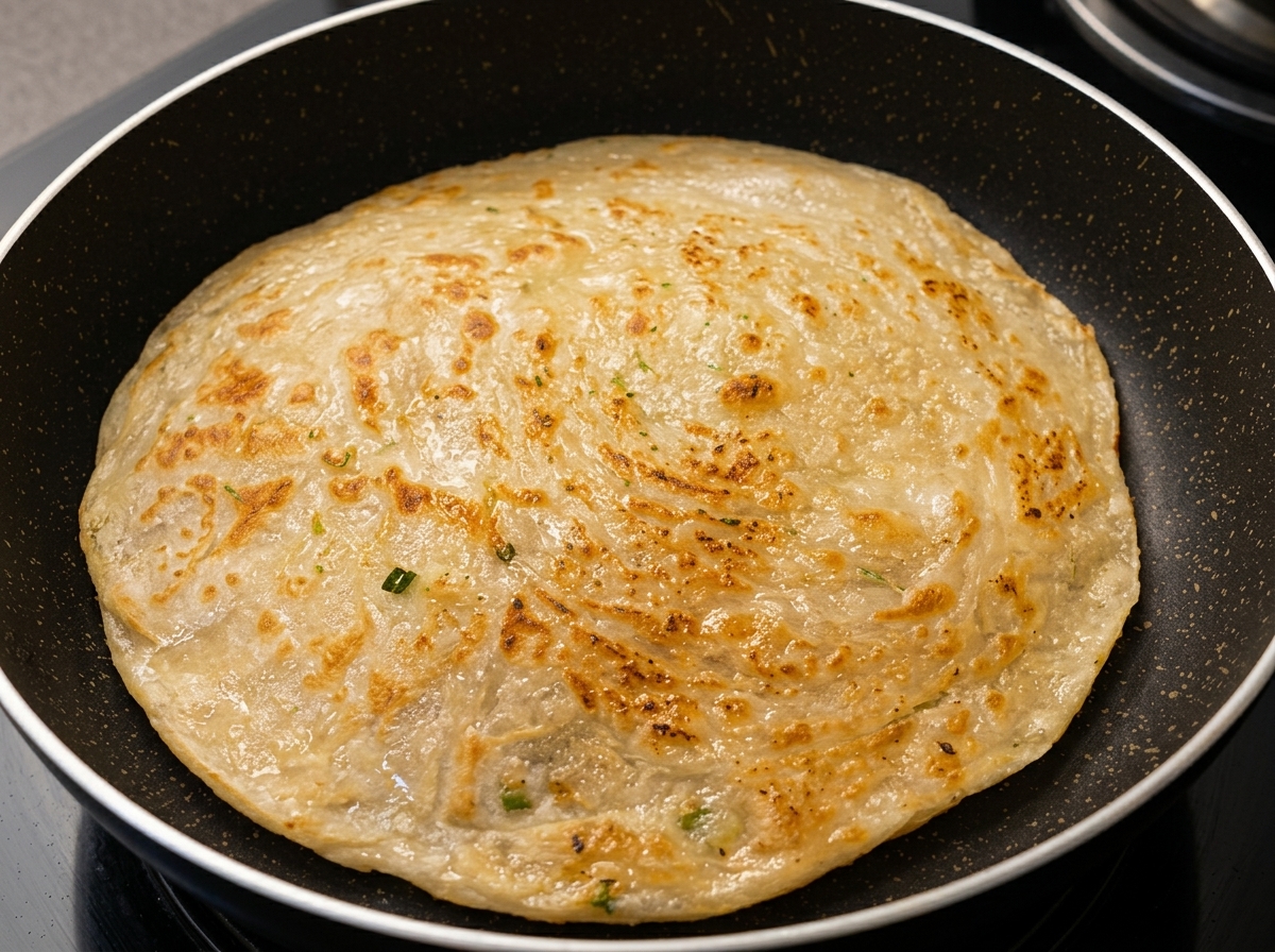 Une crêpe ronde à loignon en train de frire dans une poêle antiadhésive sombre, montrant une surface dorée et texturée.