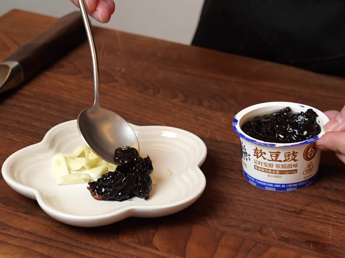 Une cuillère déposant des haricots noirs fermentés sur une petite assiette à côté de lail.