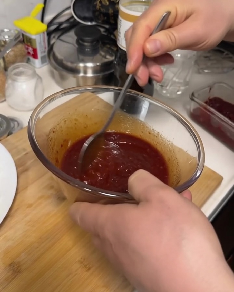 Une main utilisant une cuillère en métal pour mélanger une sauce épaisse rouge sombre dans un bol en verre transparent.