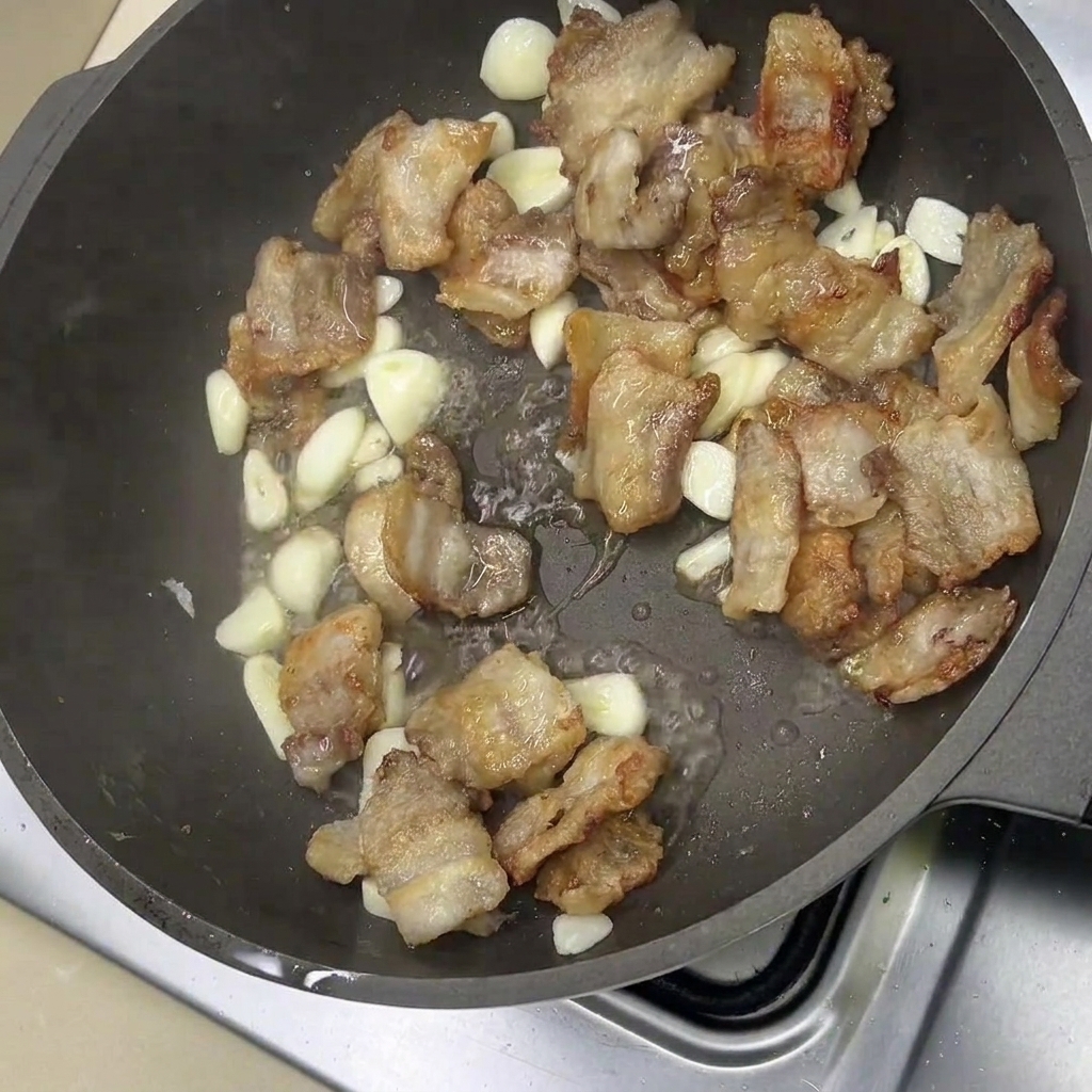 Tranches de poitrine de porc frites et gousses dail entières grésillant dans une poêle.