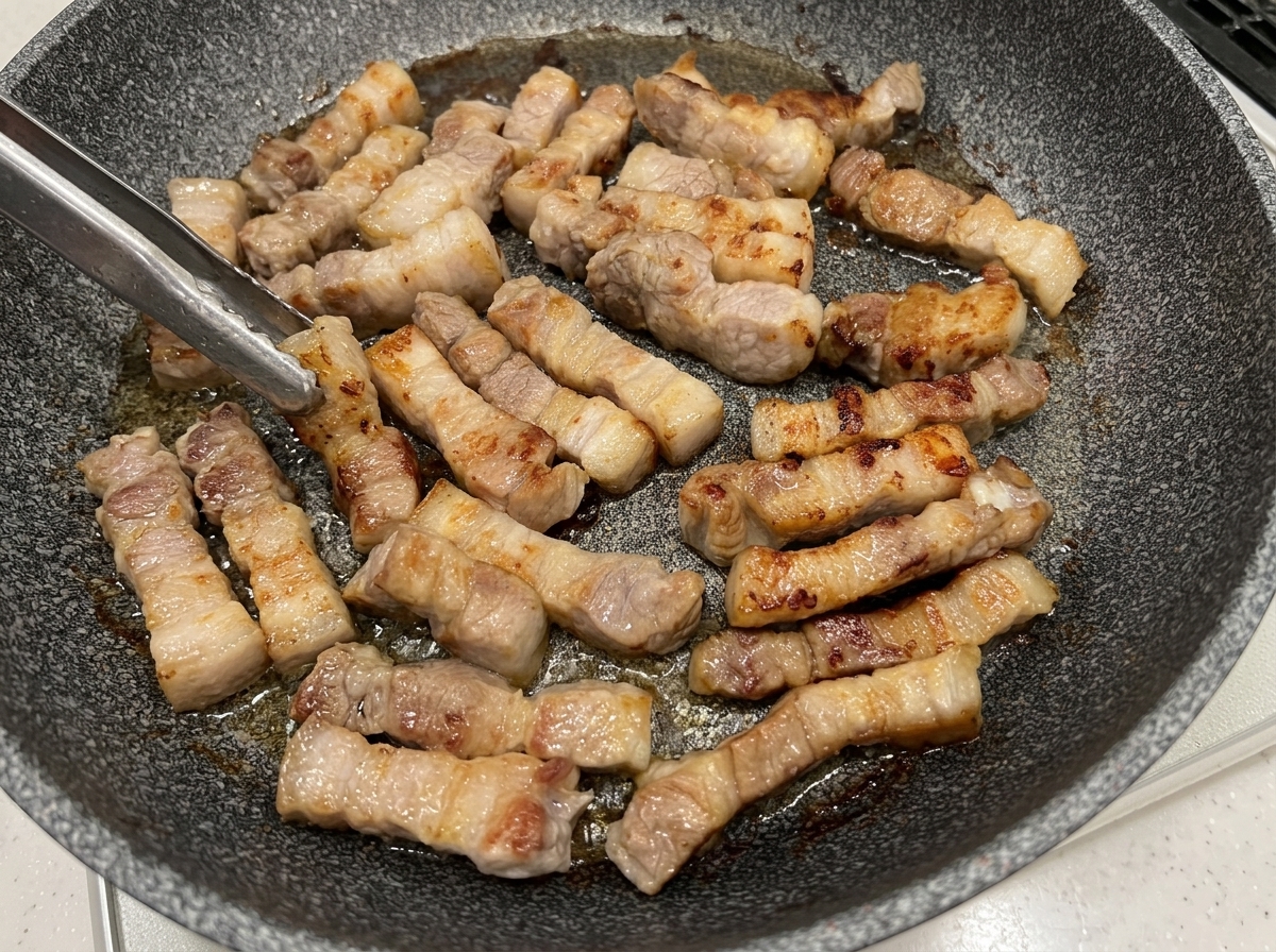 Morceaux de poitrine de porc retournés à la pince dans une poêle, montrant des surfaces dorées.