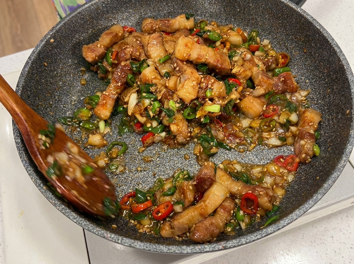 Morceaux de porc, piments et cébettes mélangés dans la poêle avec deux cuillères en bois.