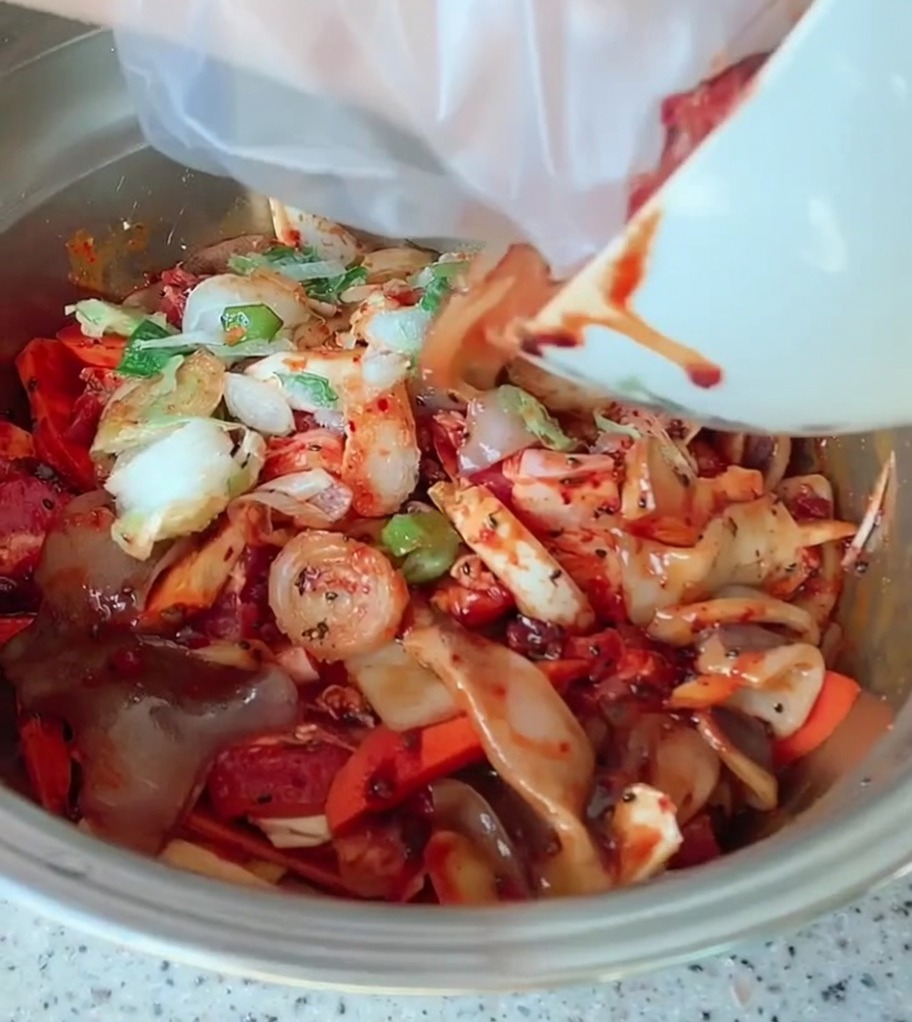 Un grand bol en métal contenant de la poitrine de porc en tranches, des légumes colorés et des oignons verts, enrobés dune sauce épicée dun rouge vif.