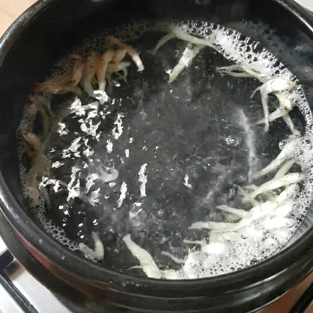 Un pot en pierre noire contenant de leau, des anchois séchés et du kelp, bouillant sur une cuisinière.