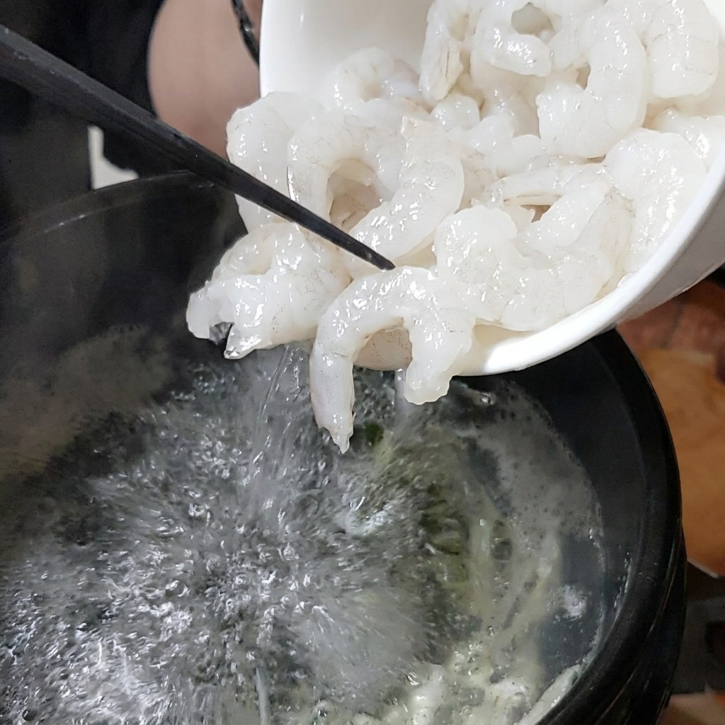 Un bol blanc versant des crevettes fraîches dans un pot de bouillon bouillonnant.