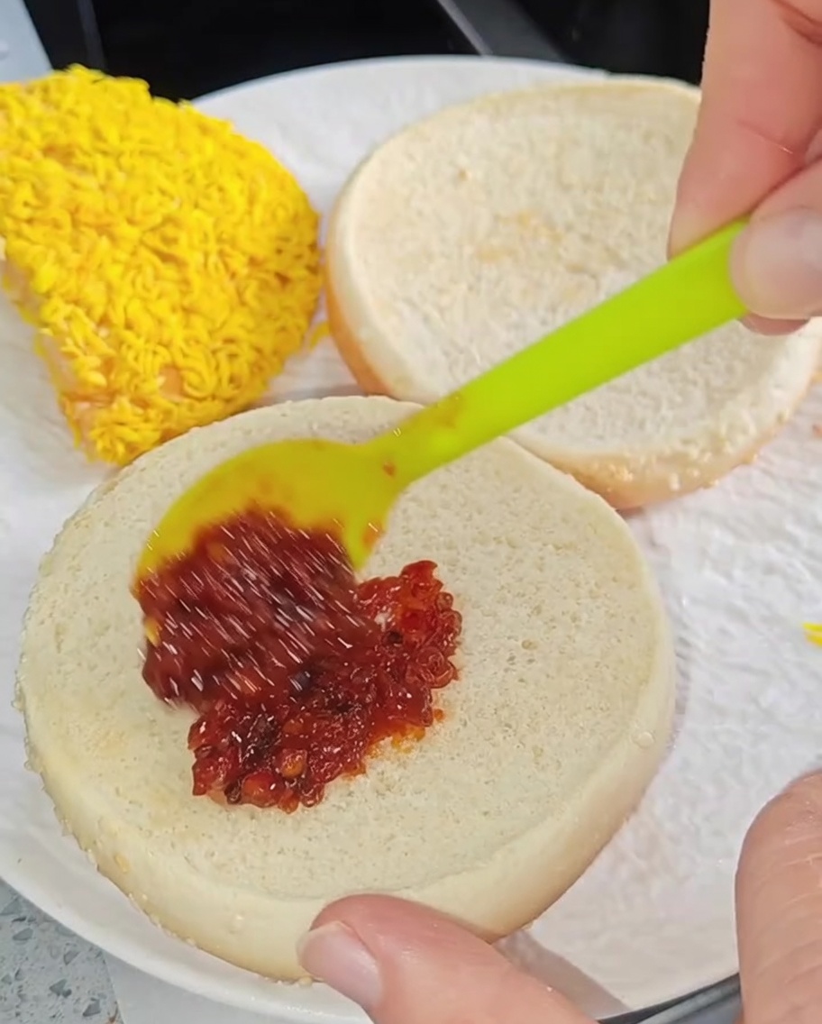 Une petite spatule étalant de la sauce chili rouge sur la face intérieure dun pain à burger grillé.
