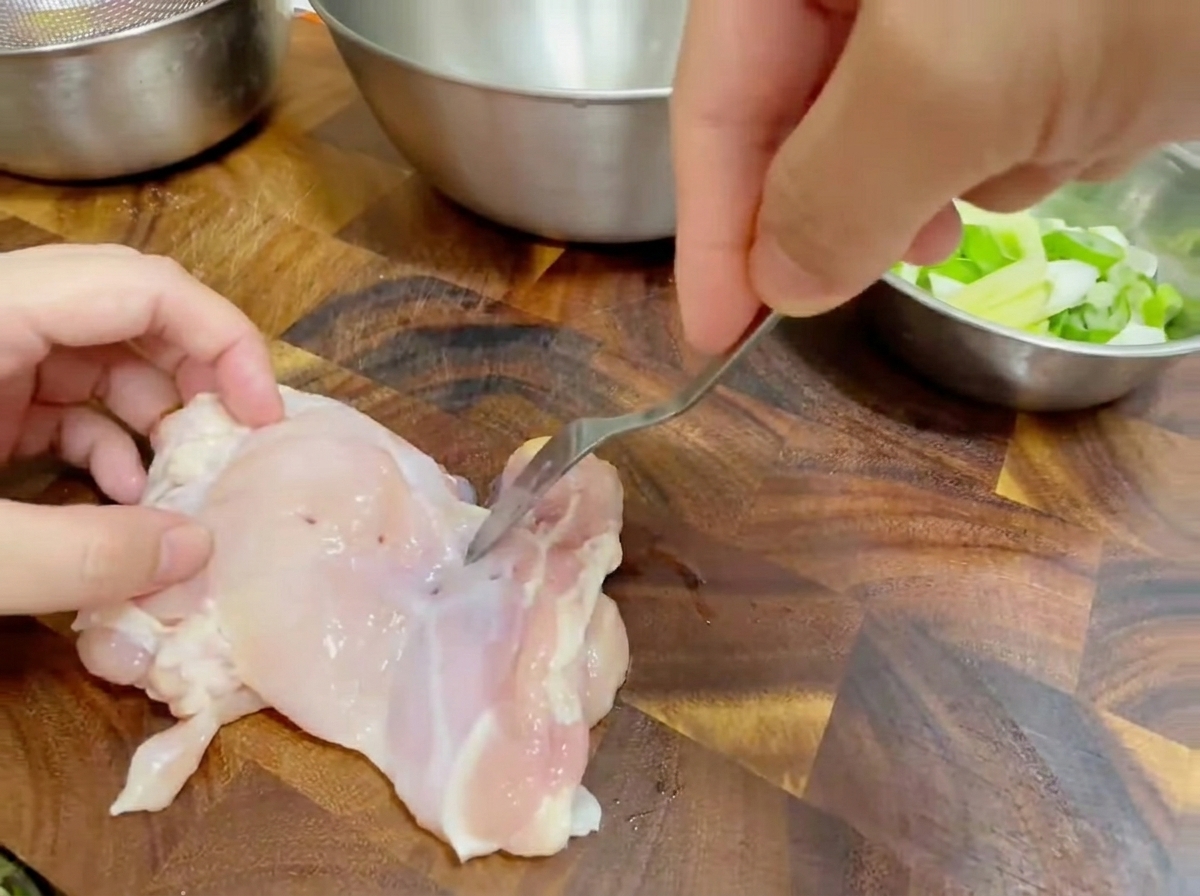 Gros plan sur une fourchette piquant une cuisse de poulet crue désossée sur une planche à découper en bois.