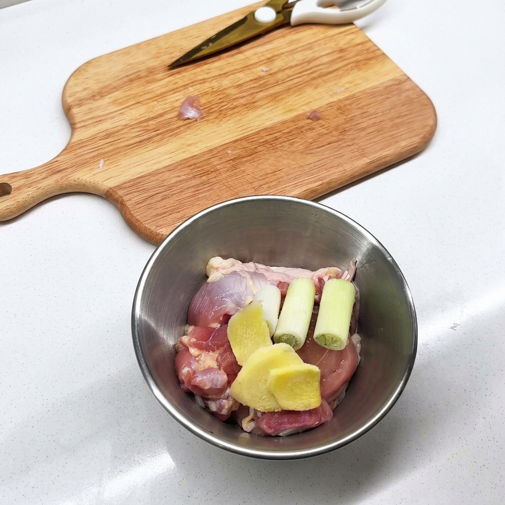 Bol en métal contenant des morceaux de poulet cru, des tranches de gingembre et de loignon vert.