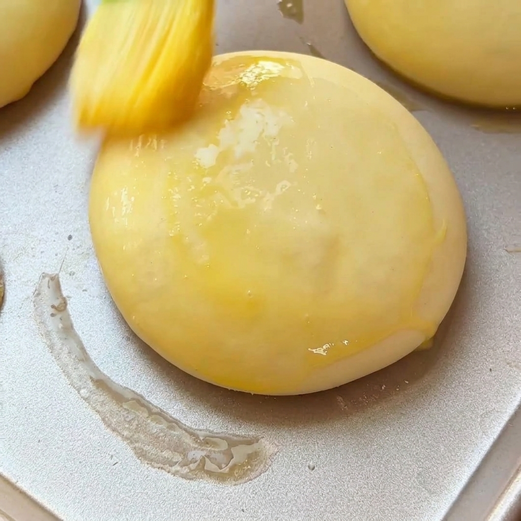 Un pinceau à pâtisserie badigeonnant de dorure un pain à burger levé sur une plaque de cuisson en métal.