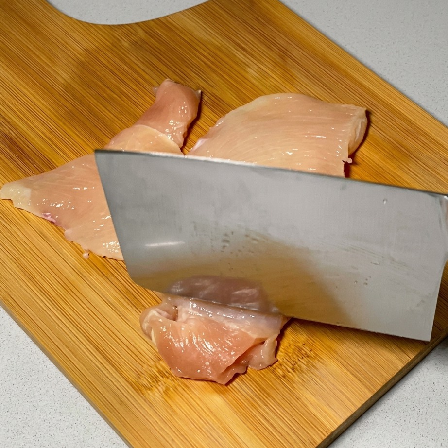 Un couteau de cuisine utilisé pour attendrir des tranches de poulet cru sur une surface en bois.