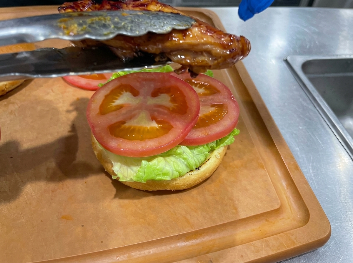 Une cuisse de poulet teriyaki cuite déposée avec des pinces sur un burger monté avec salade et tomate.
