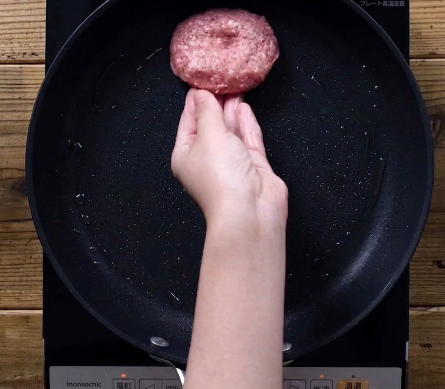 Une main déposant délicatement une épaisse galette de viande crue dans une poêle noire sur une plaque à induction.