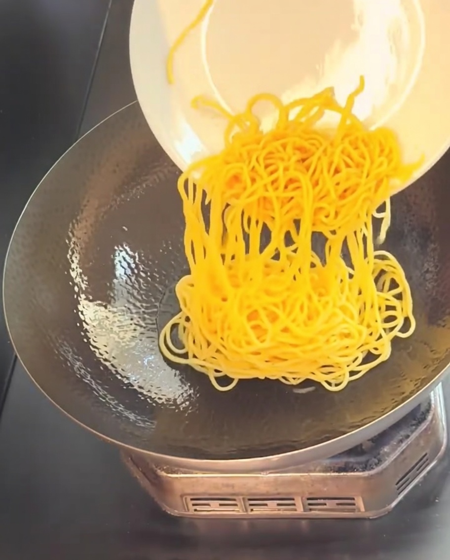 Des spaghettis jaunes cuits glissés dune assiette blanche dans un wok chaud et huilé.