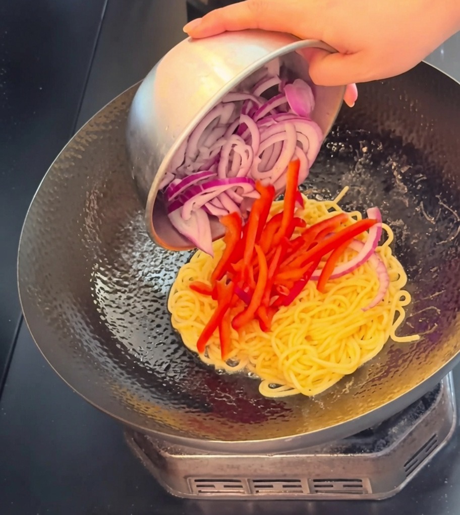 Vue rapprochée de lamelles de bœuf, de lanières de poivron rouge et doignon rouge ajoutées dans un wok contenant les spaghettis.