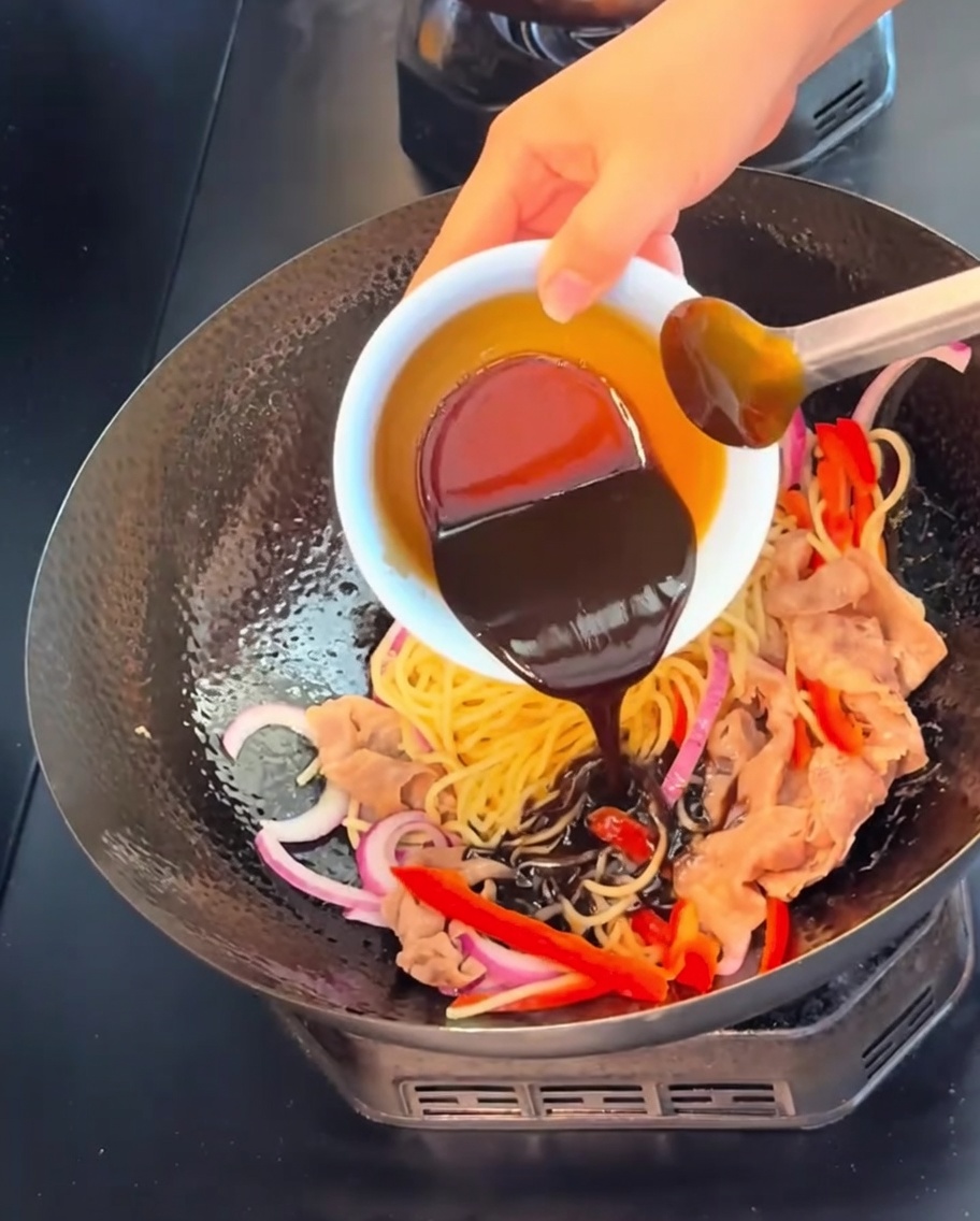 Une sauce au poivre noir riche et foncée versée dun petit bol blanc sur les nouilles, le bœuf et les légumes dans un wok.