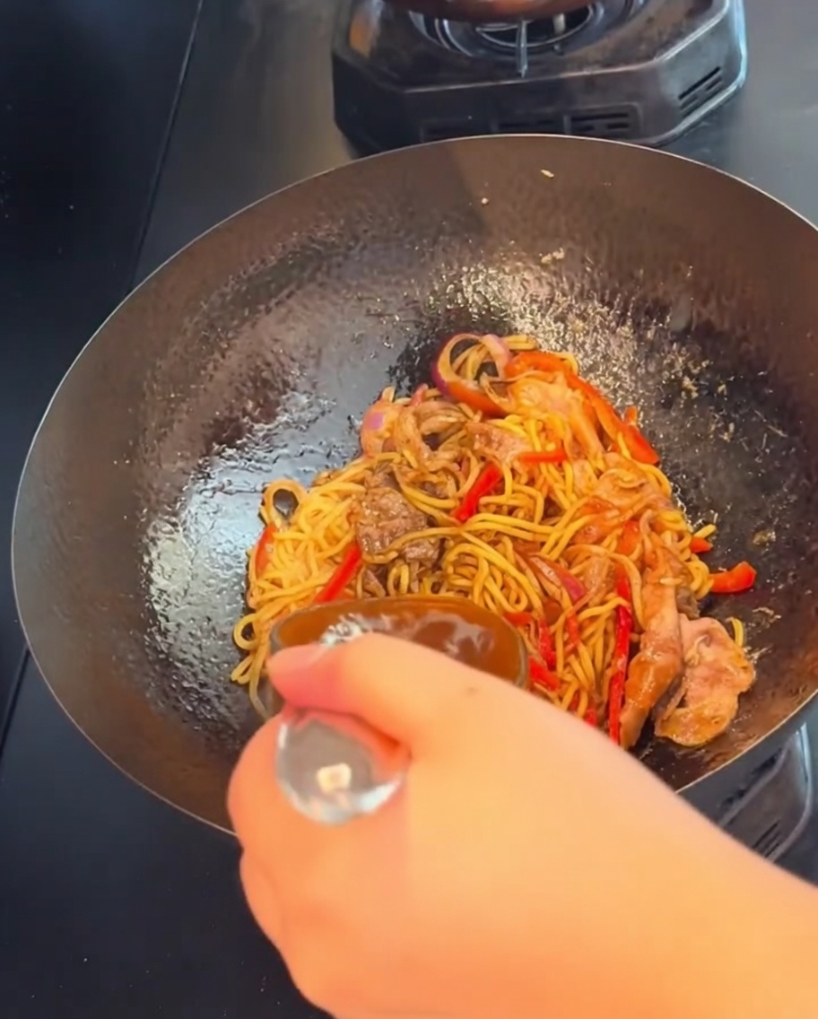 Une main tenant une spatule, faisant sauter les spaghettis au bœuf au poivre noir à feu vif dans un wok.