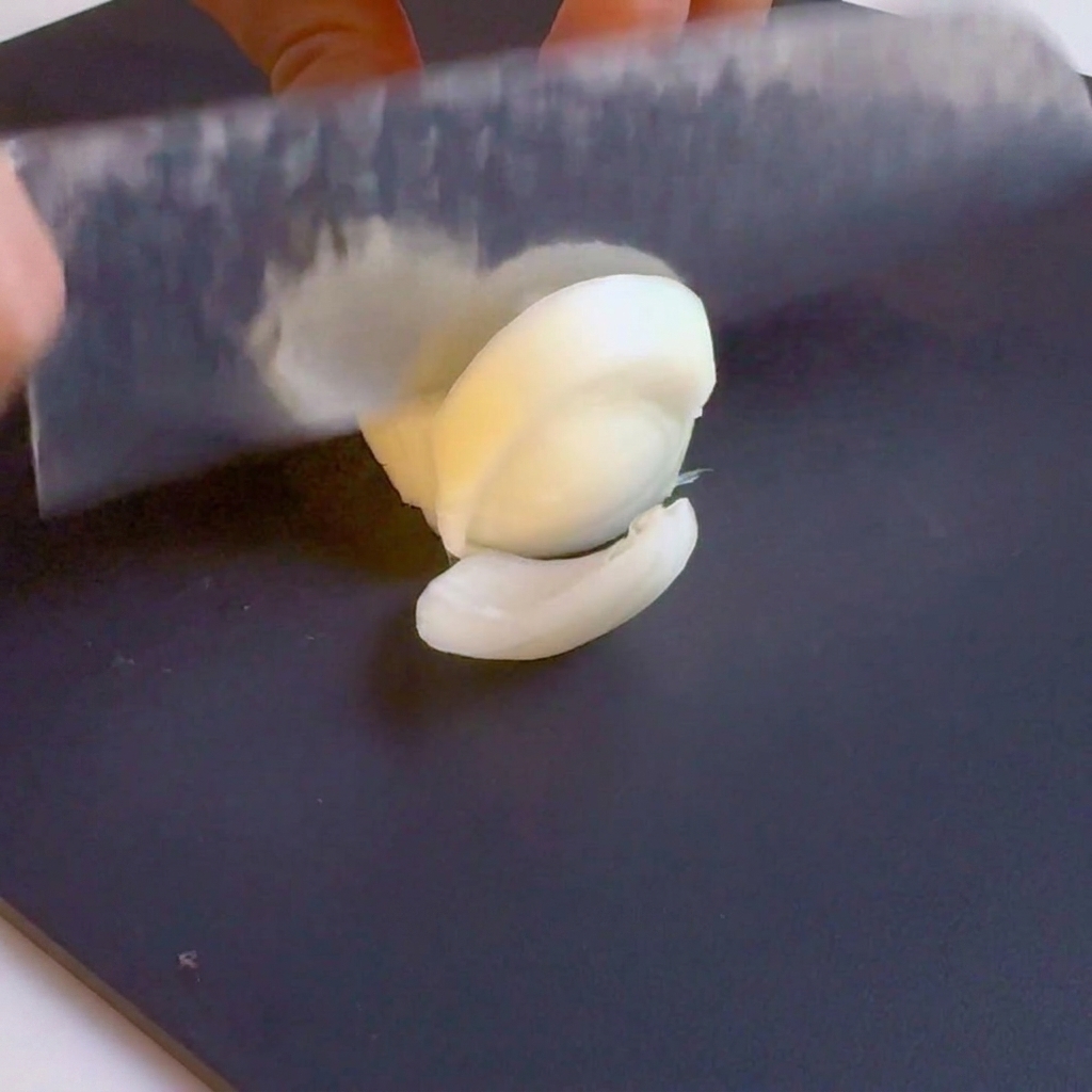 Un couteau éminçant un oignon blanc en lamelles sur une planche à découper noire.