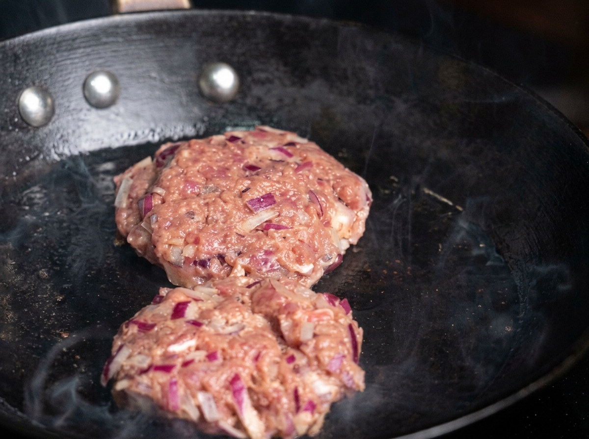 Deux steaks de bœuf assaisonnés déposés dans une poêle chaude huilée, avec des volutes de fumée visibles.