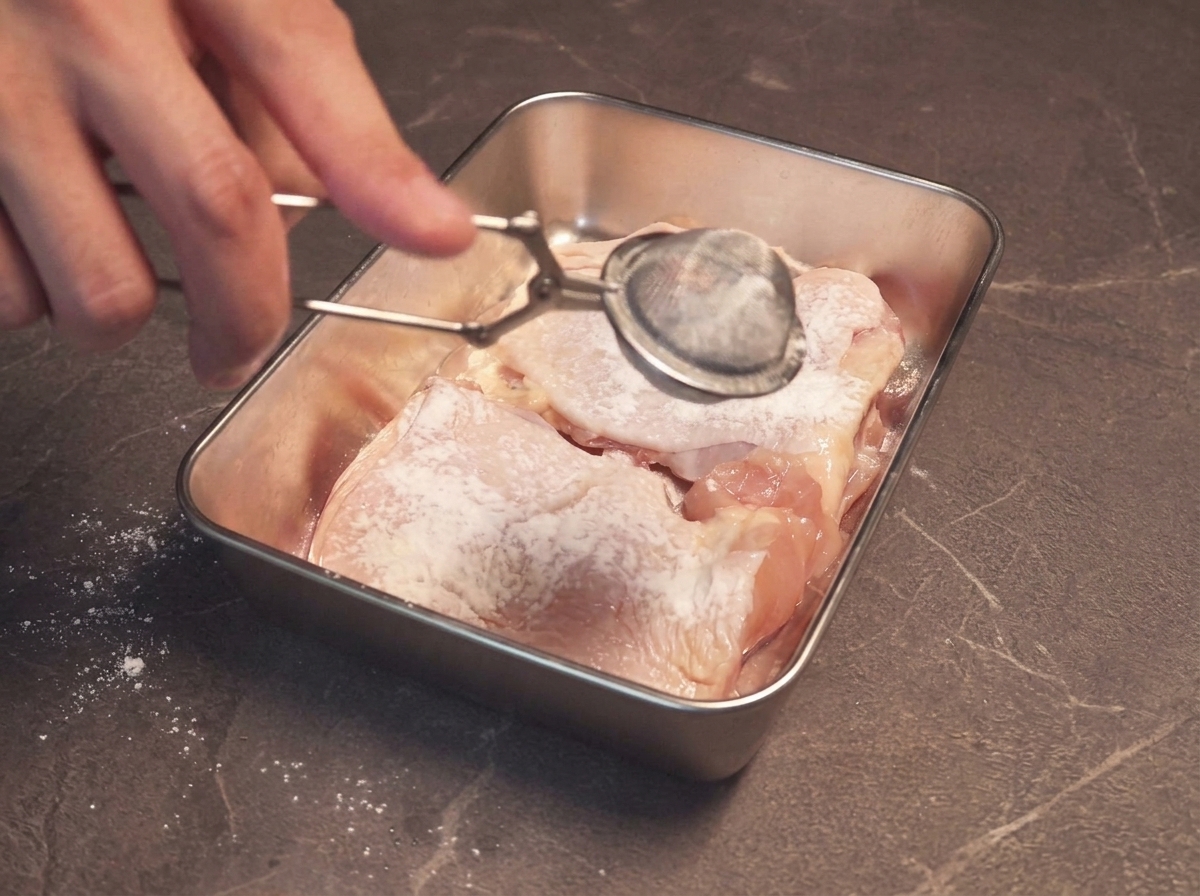 Fécule saupoudrée à laide dune passoire fine sur les morceaux de poulet marinés disposés dans un plateau en inox.