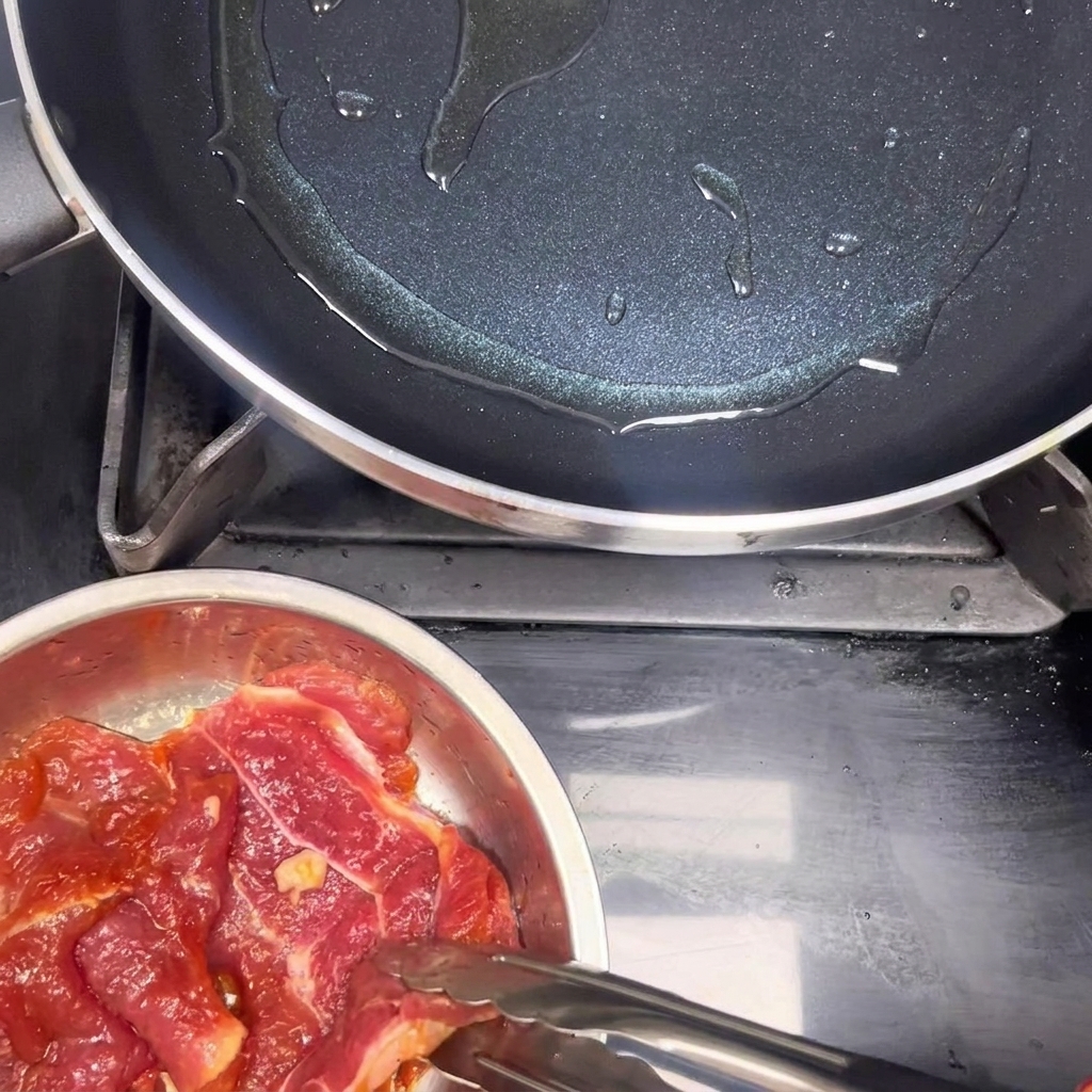 Des pinces de cuisine déposent des morceaux de porc cru mariné dans une poêle antiadhésive chaude huilée.