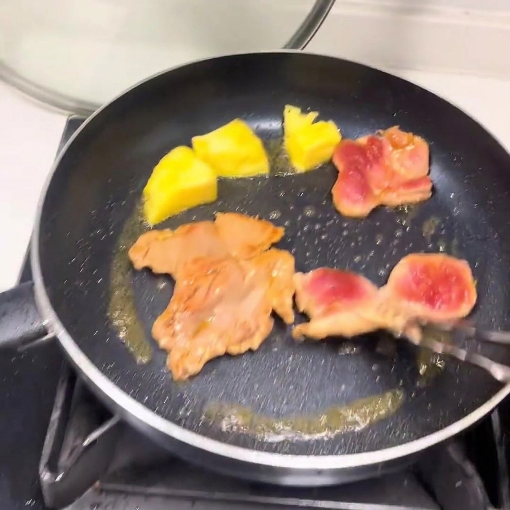 Tranches de porc dorées et morceaux dananas en train de cuire sur leur seconde face dans une poêle.
