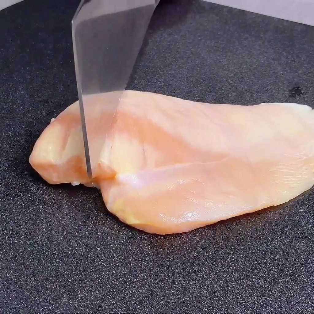 Aplatir un blanc de poulet cru avec le dos dun grand couteau sur une planche à découper foncée.