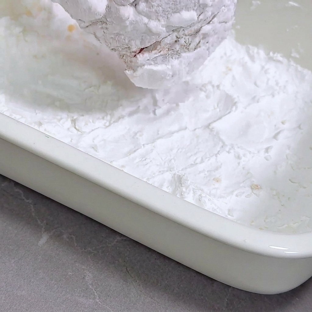 Un blanc de poulet mariné pressé dans un plateau rempli de fécule de maïs.