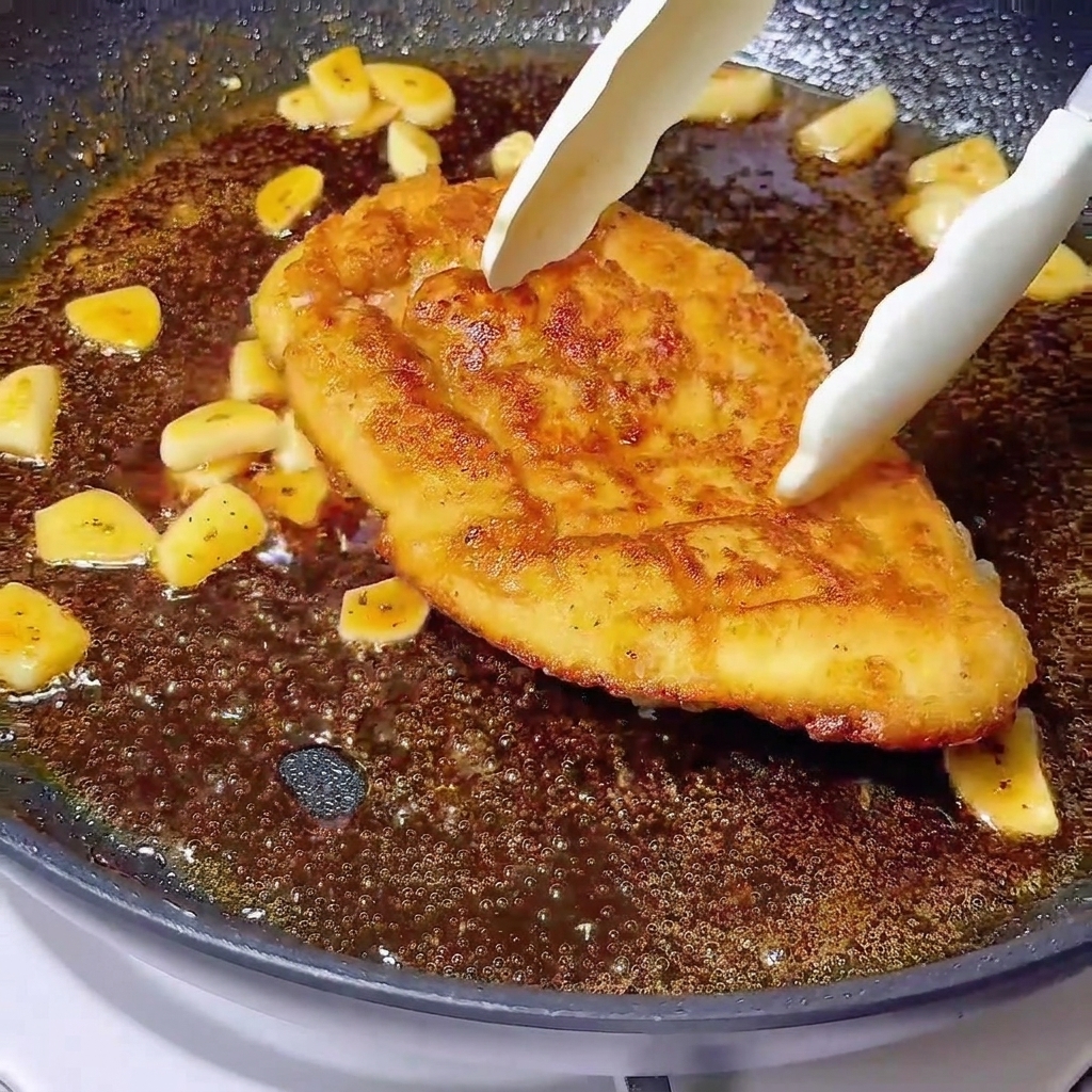 Des pinces déposant un blanc de poulet doré dans un glaçage bouillonnant avec de lail.
