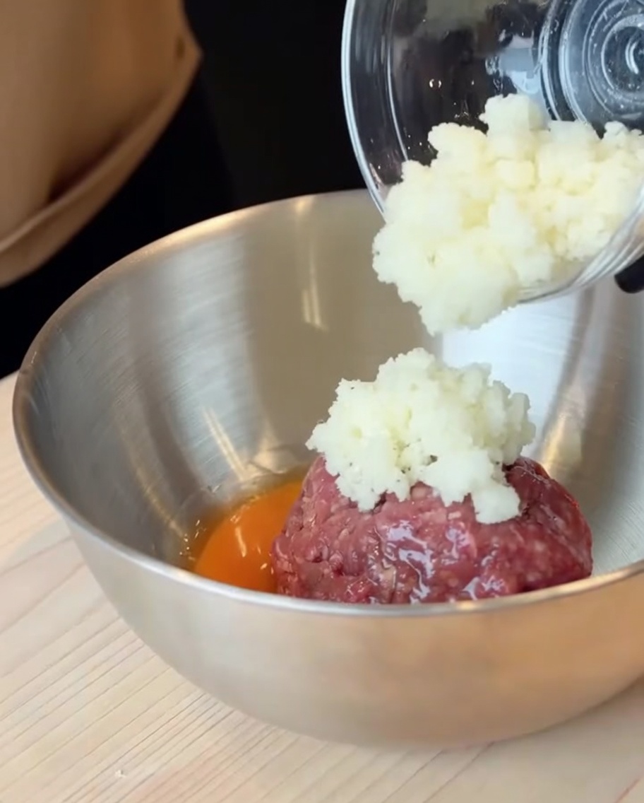 Bœuf haché cru et un œuf cassé dans un grand bol en inox avec loignon ajouté.