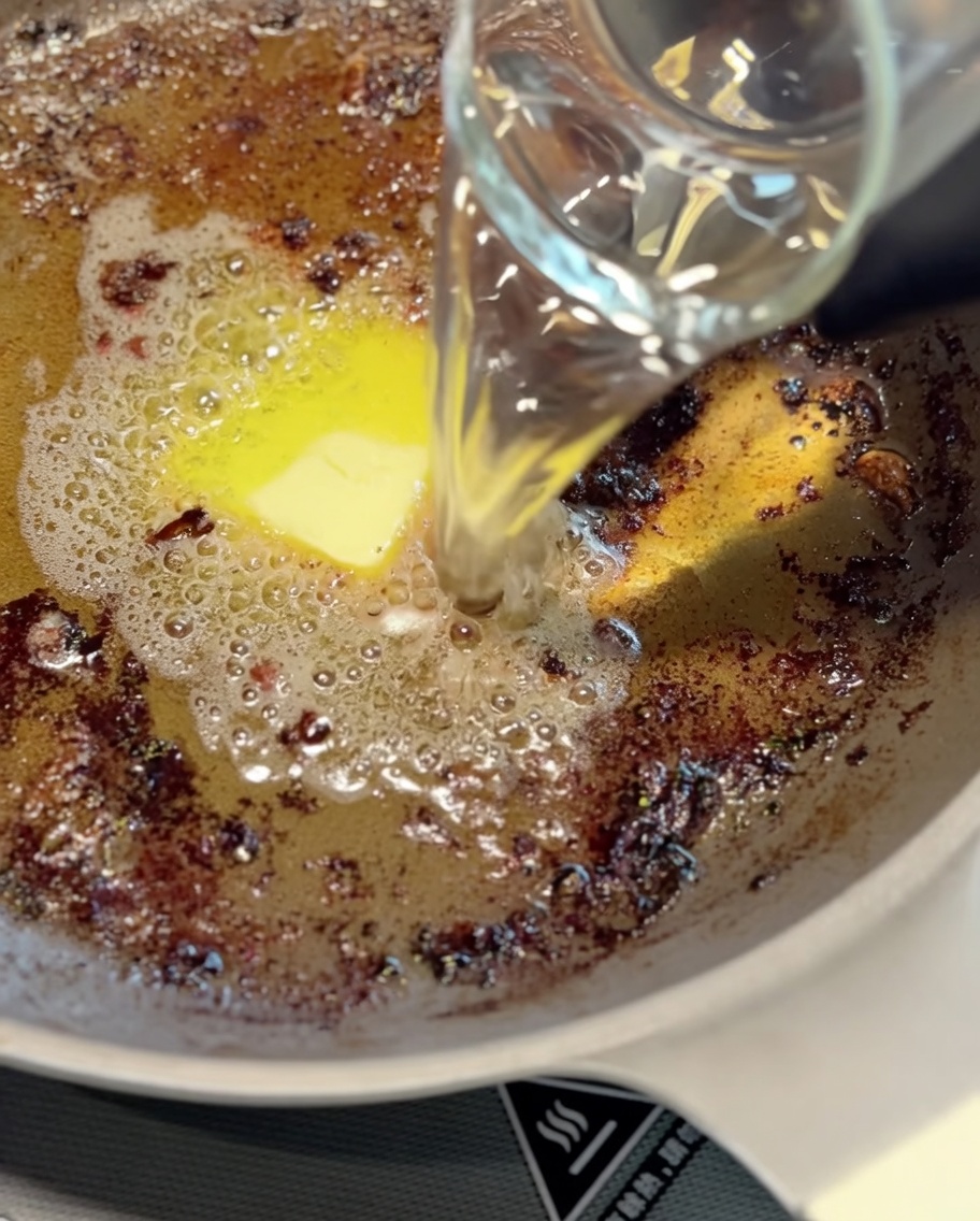 Une louche en métal remuant les jus de cuisson et leau dans une poêle pour former la base de la sauce.