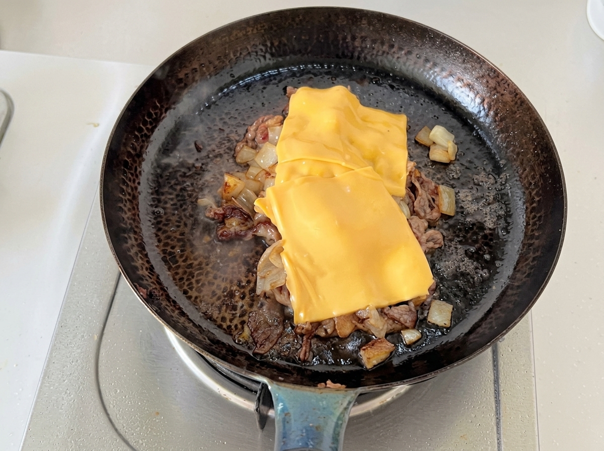 Deux tranches de fromage jaune fondant sur du bœuf émincé et des oignons cuits dans une poêle.
