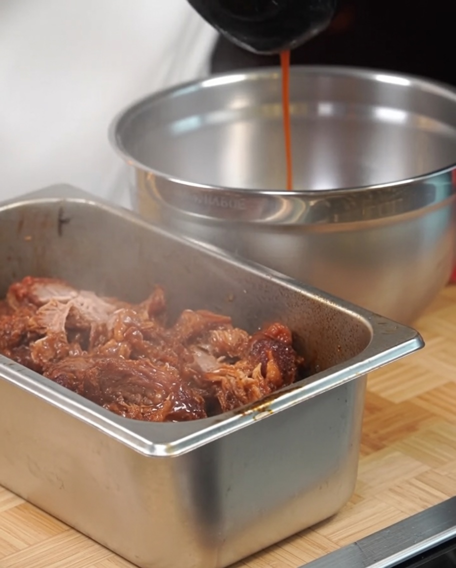 Porc effiloché dans un plat métallique assaisonné avec du jus de cuisson à laide dune louche.