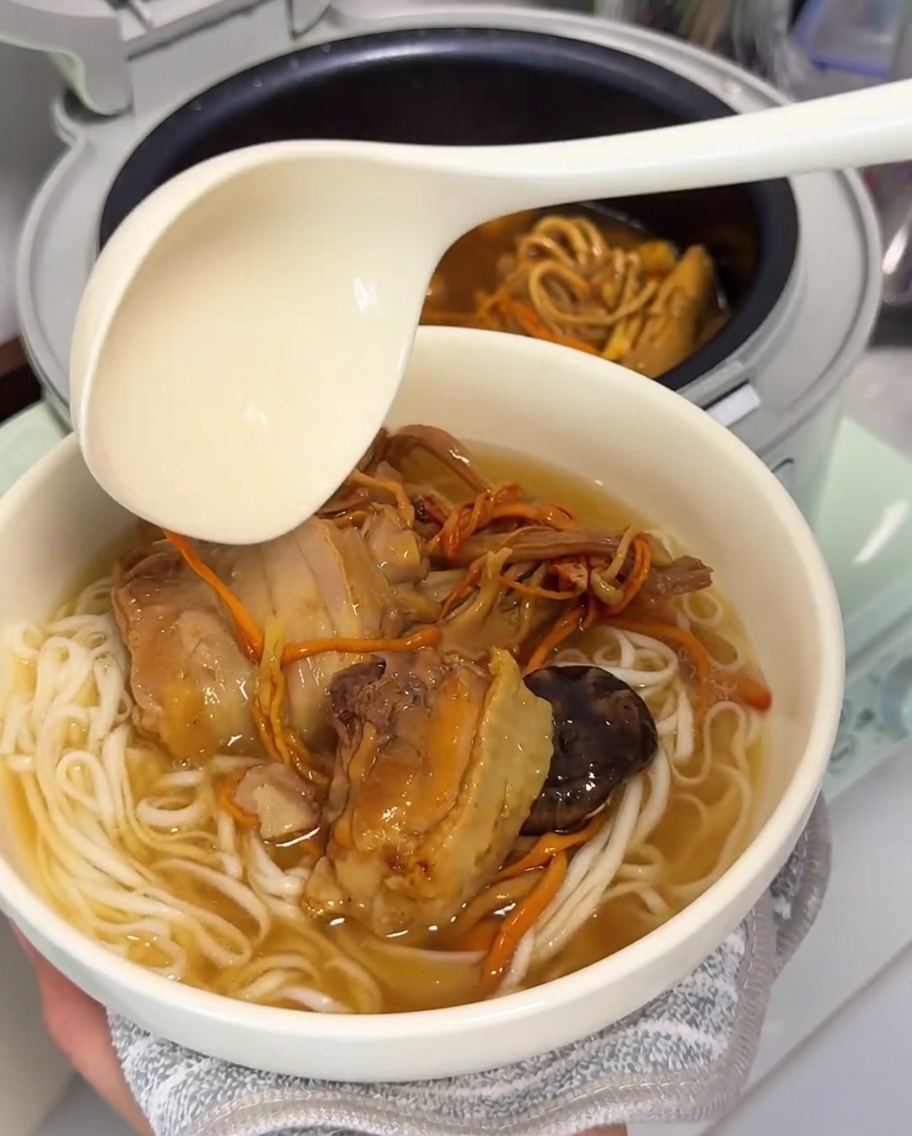 Une grande louche versant le bouillon doré de poulet et champignons sur un nid de nouilles fraîchement cuites.