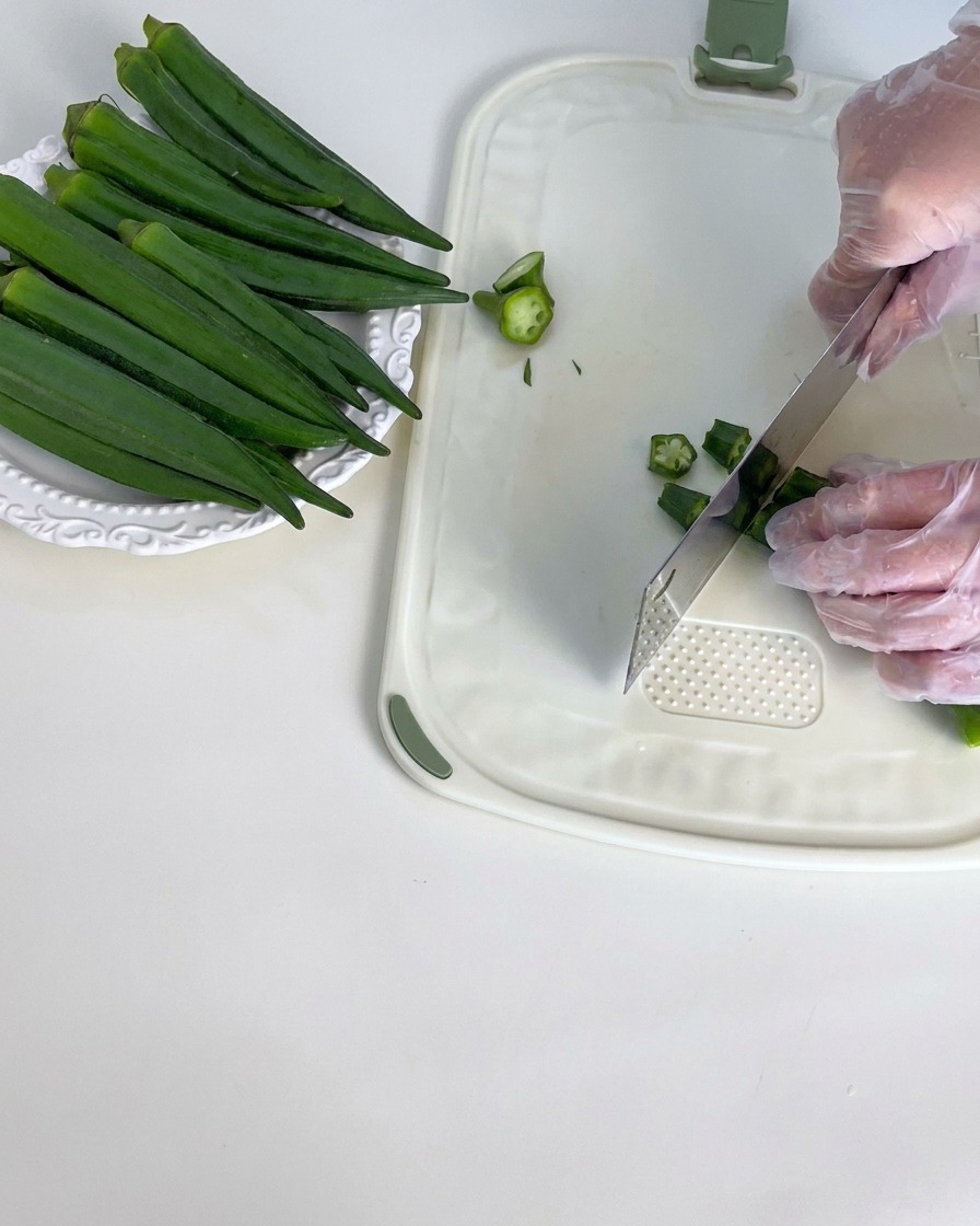 Gousses de gombo vert frais en cours déqueutage sur une planche à découper blanche.