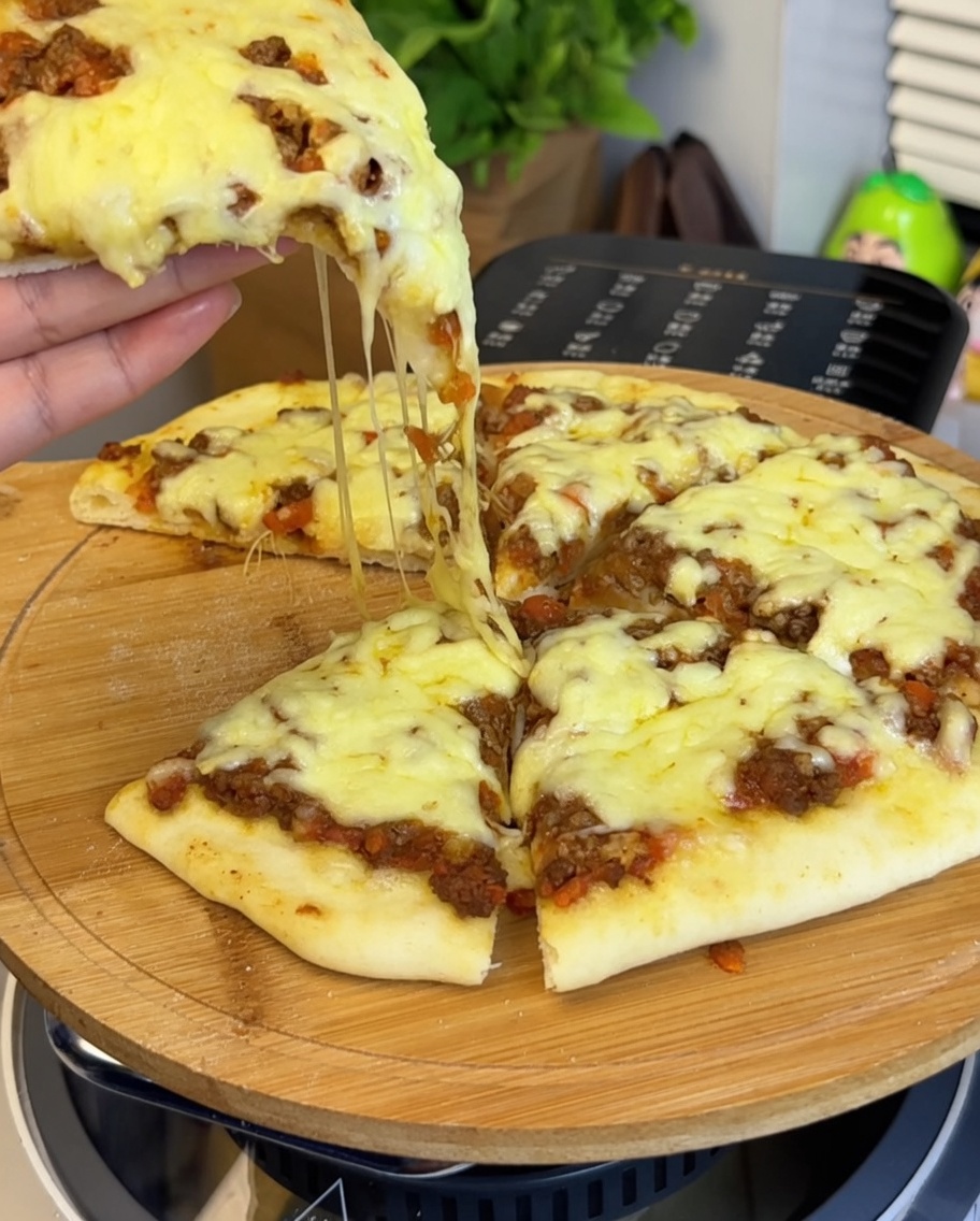 Gros plan sur la sauce tomate au bœuf étalée sur la pâte à pizza, avec une main tenant une part de pizza au premier plan montrant le fromage fondu.