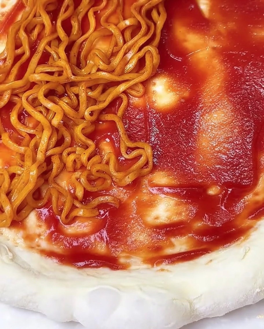 Gros plan sur une pâte à pizza nappée de sauce tomate, partiellement recouverte de nouilles ramen épicées et de paillettes dalgues.