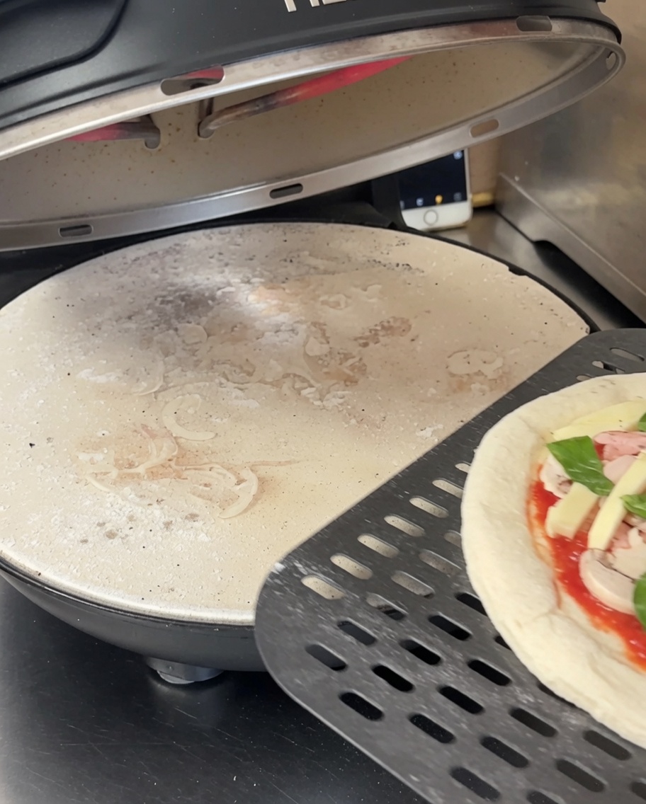 Pâte à pizza étalée sur une pierre à pizza noire perforée à lintérieur dun four à pizza.