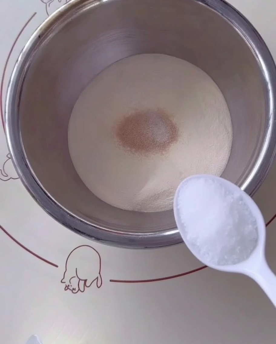 Une cuillère de sucre ajoutée au mélange de farine et de levure dans un bol en acier.