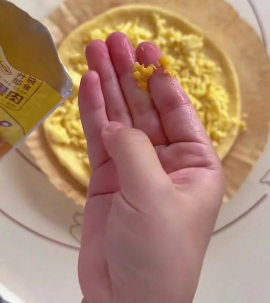 Une main dispersant des grains de maïs jaunes sur une moitié de la base de pâte à pizza ronde.